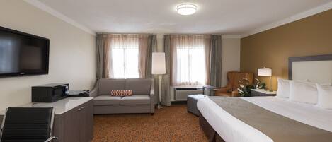 Habitación de hotel con cama, sofá, escritorio con computadora, televisor y ventana con cortinas.