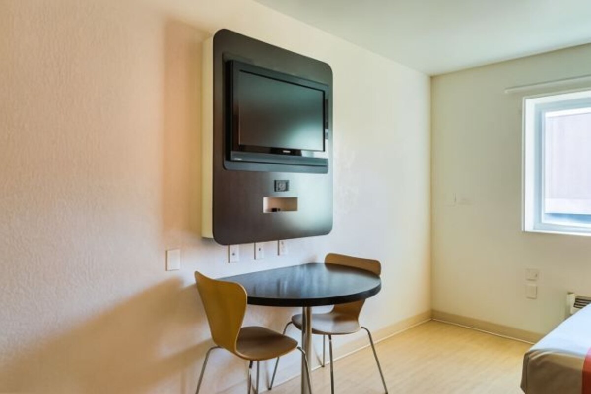 A room with a flat-screen TV mounted on the wall, a round table with two chairs, and a window providing natural light.
