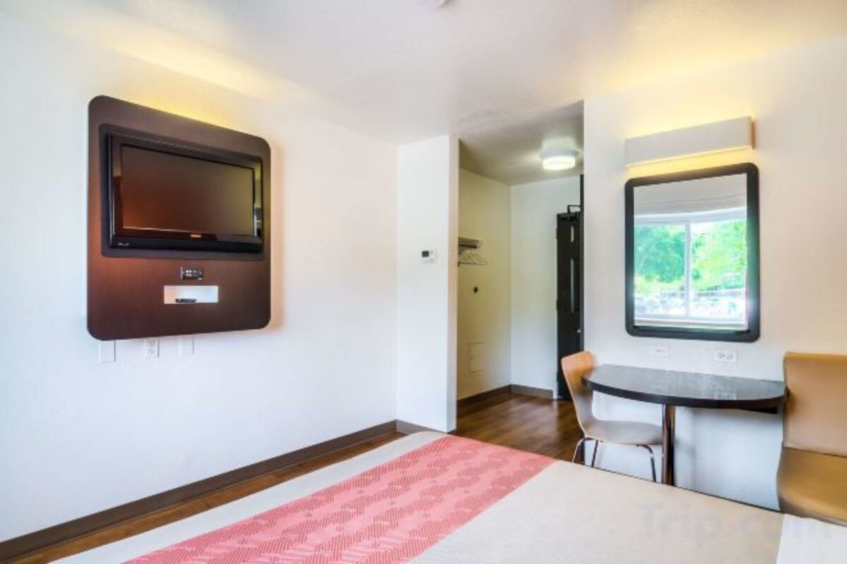 A hotel room with a flat-screen TV mounted on the wall, a small table with a chair, and a window with a view of greenery.