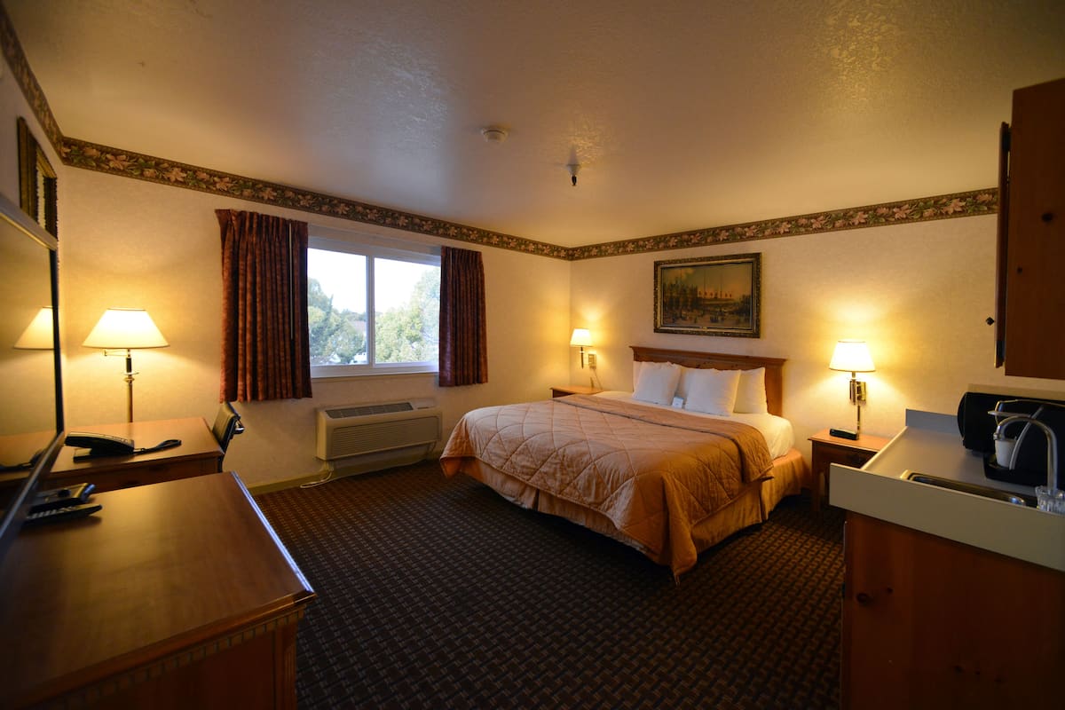 A hotel room with a bed, bedside lamps, a desk with a telephone, a window with curtains, and a painting on the wall.
