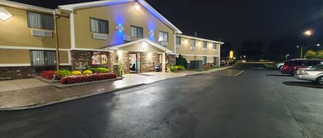 A hotel with a well-lit entrance, a parking area, and a landscaped front garden.