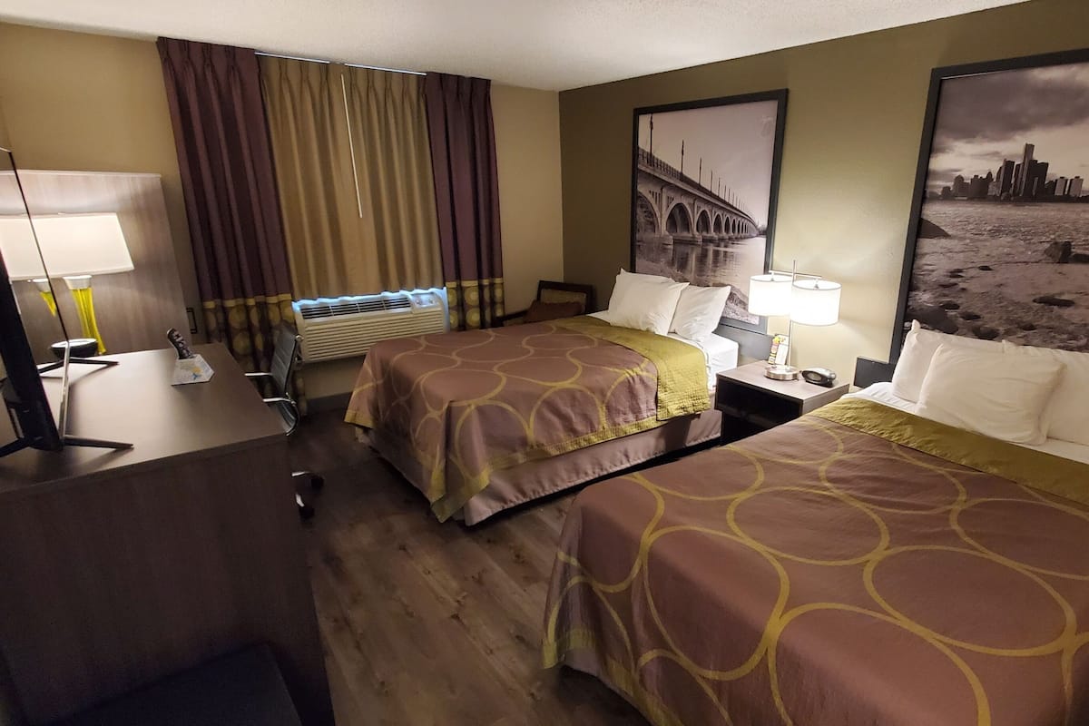 Ett hotellrum med två sängar, ett skrivbord med en lampa, en luftkonditionering och en stor tavla med en stadsbild på väggen.