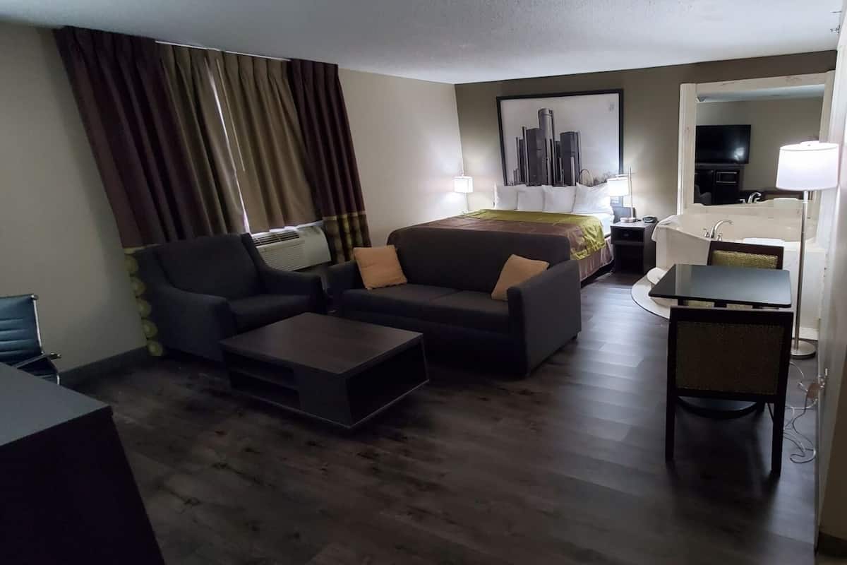 Ett hotellrum med ett vardagsrumsutrymme som inkluderar en soffa, ett soffbord och en TV. Det finns ett litet bord med en lampa och en säng med ett grönt överkast.