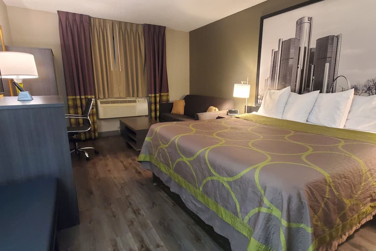 Ett hotellrum med en säng, ett skrivbord med en lampa, en stol, en luftkonditioneringsanläggning och en stor målning av en stadsbild på väggen.