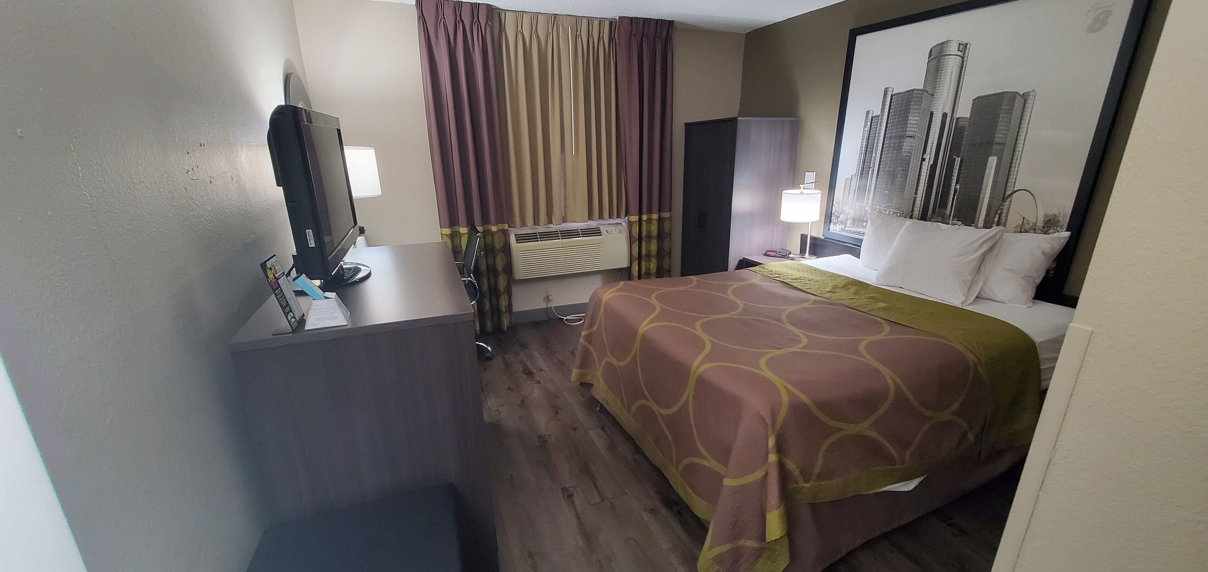 A hotel room with a bed, a desk with a computer, a window with curtains, a wall-mounted air conditioner, and a cityscape painting.