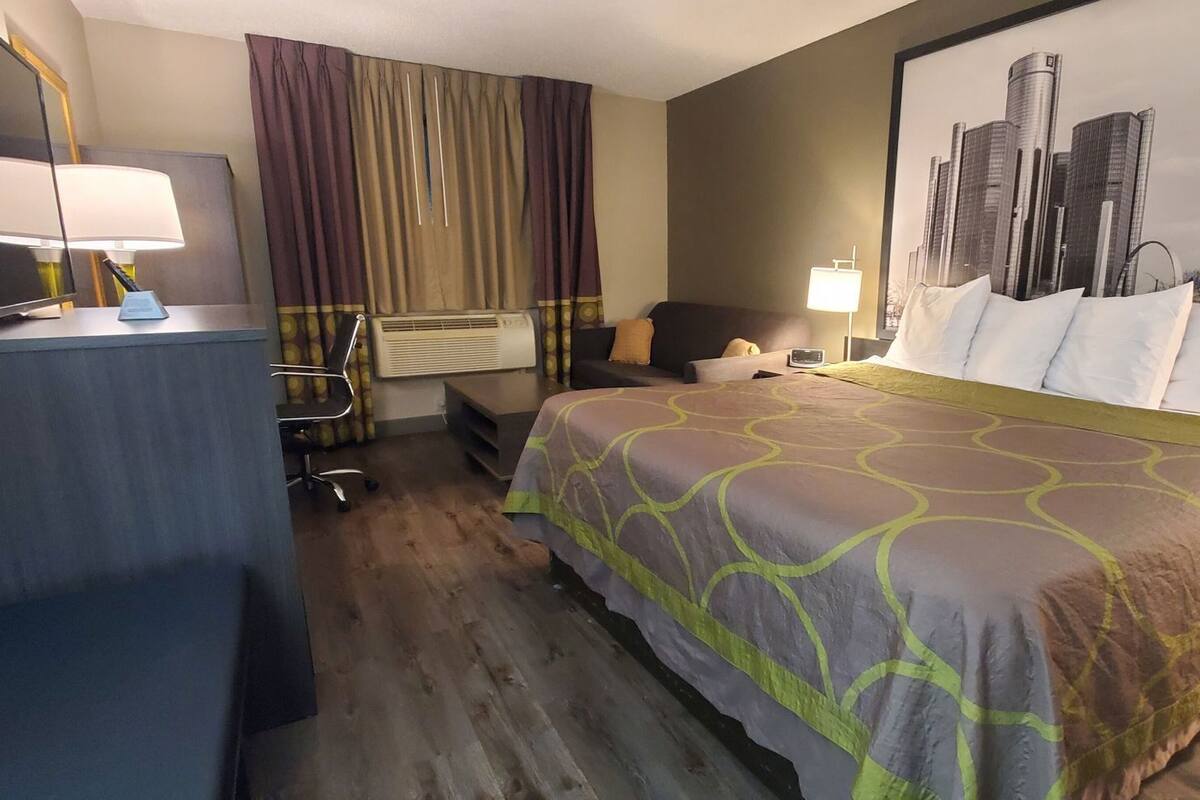 Ett hotellrum med en säng, ett skrivbord med en lampa, en stol, ett fönster med gardiner och en tavla med en stadsbild på väggen.
