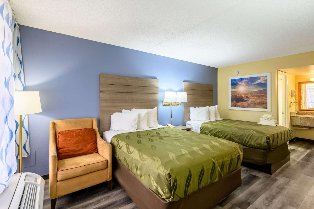 A hotel room with two beds, a chair, a lamp, and a painting on the wall.