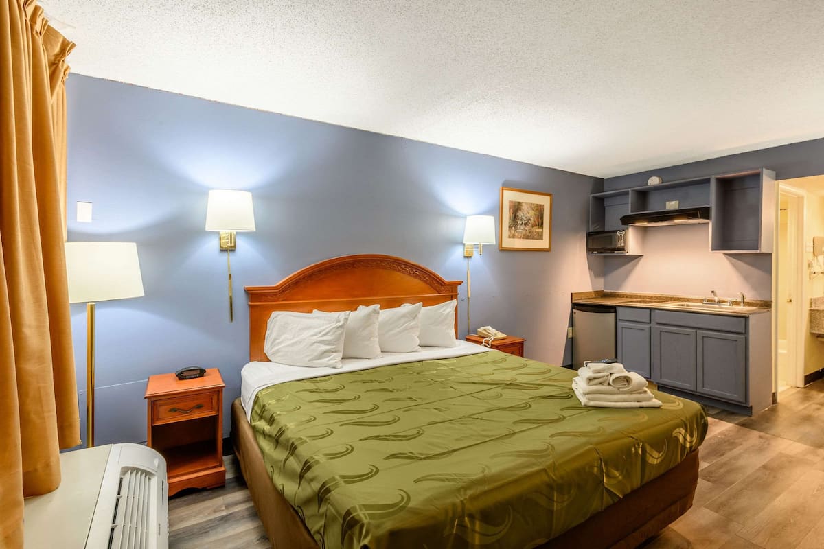 A hotel room with a wooden bed, a nightstand, a desk with a telephone, a kitchenette, and a bathroom.