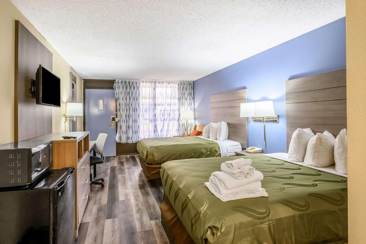 A hotel room with two beds, a desk with a computer, a TV, and a window with patterned curtains.