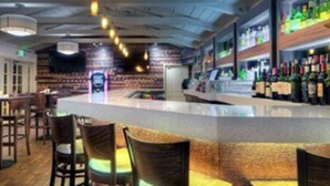 Eine Bar mit Holzsesseln, einer Theke mit Präsentationsfläche und Regalen mit Flaschen.