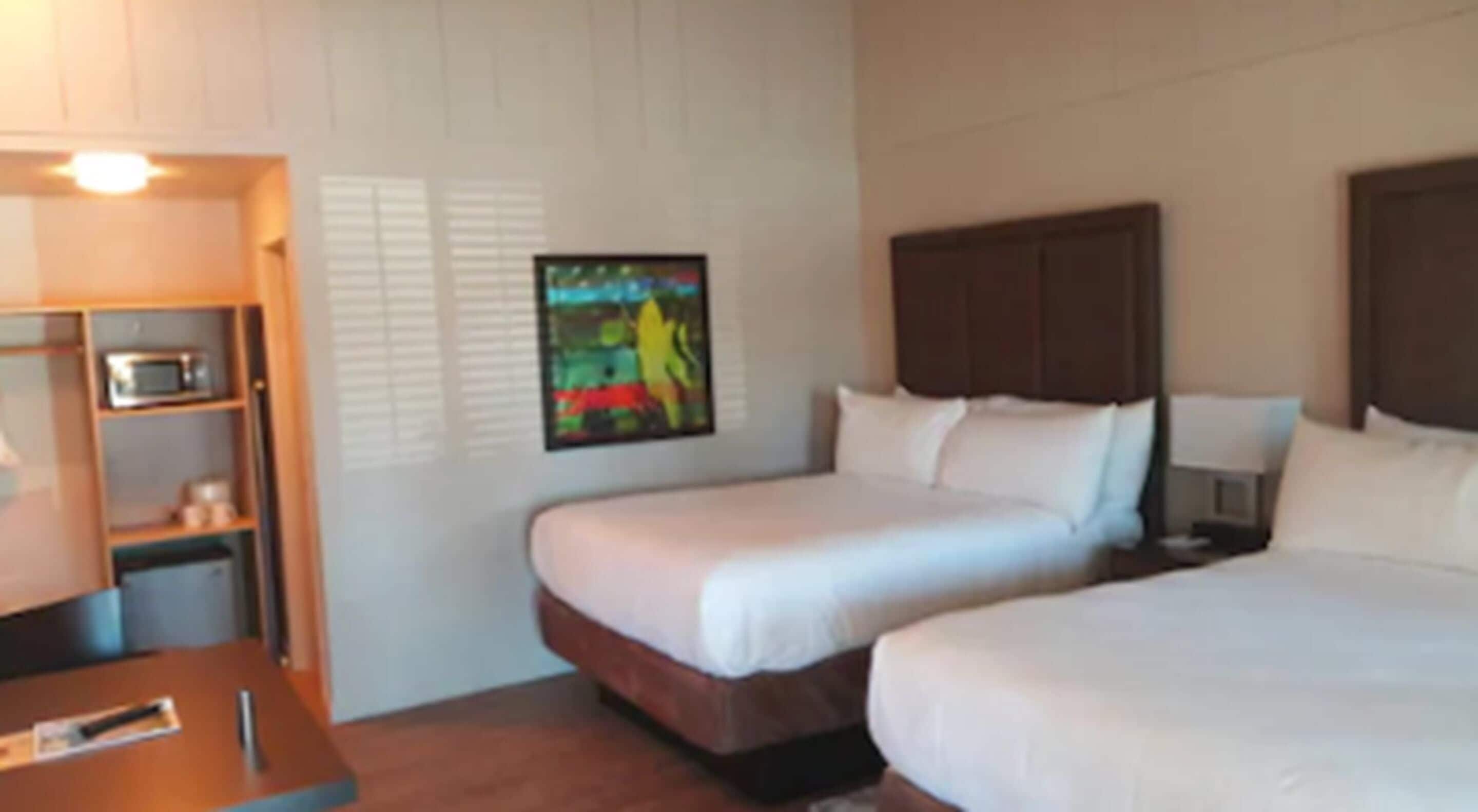 A hotel room with two beds, a desk, a microwave, and a painting on the wall.