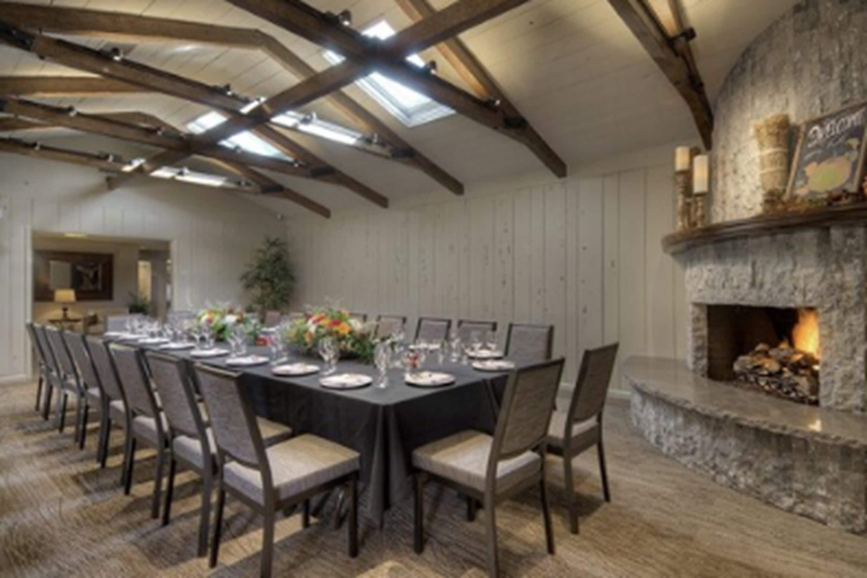 A dining area with a long table set for a meal, wooden beams on the ceiling, and a stone fireplace.