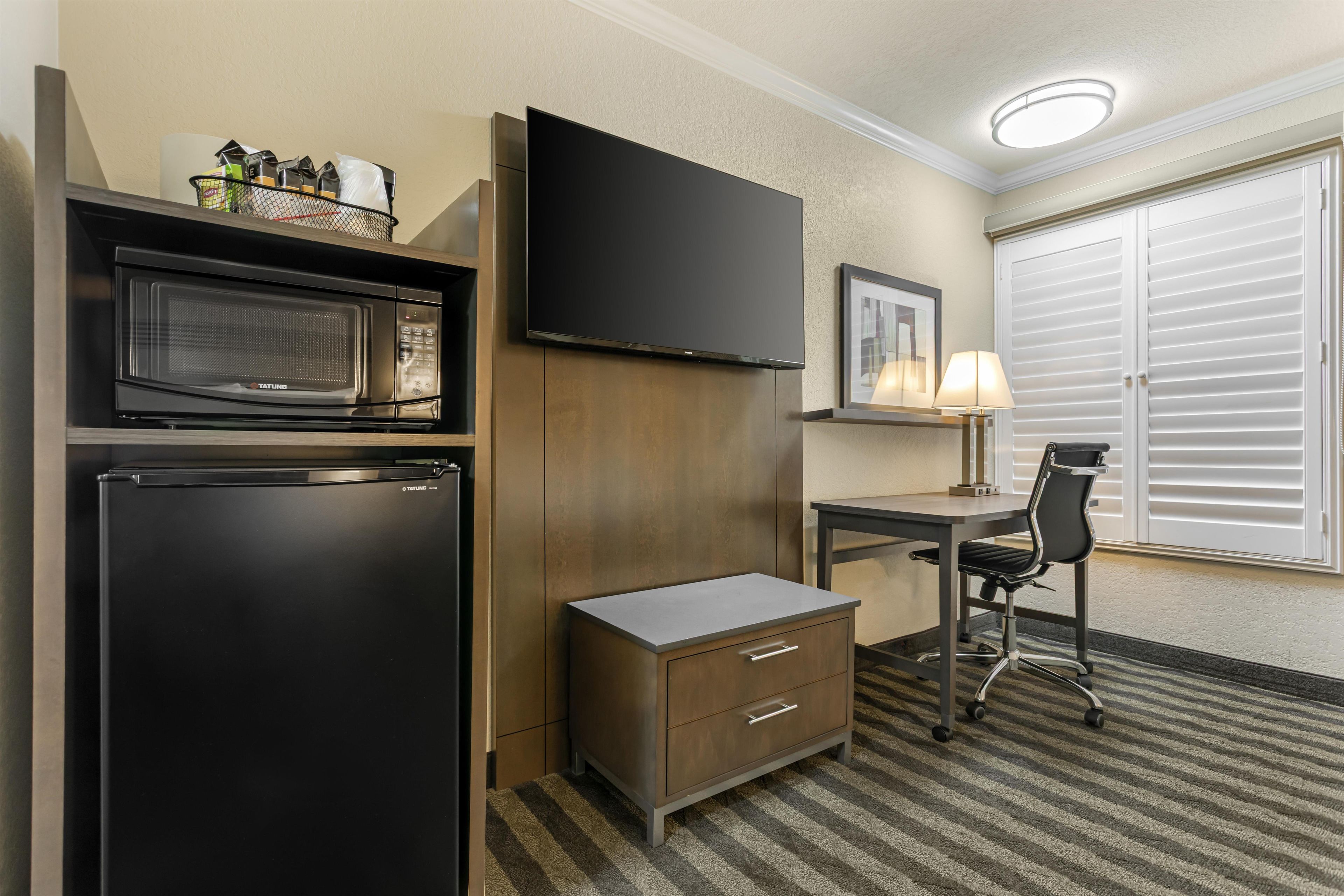 Habitación de hotel con kitchenette, televisión de pantalla plana, escritorio con silla y ventana con persianas.