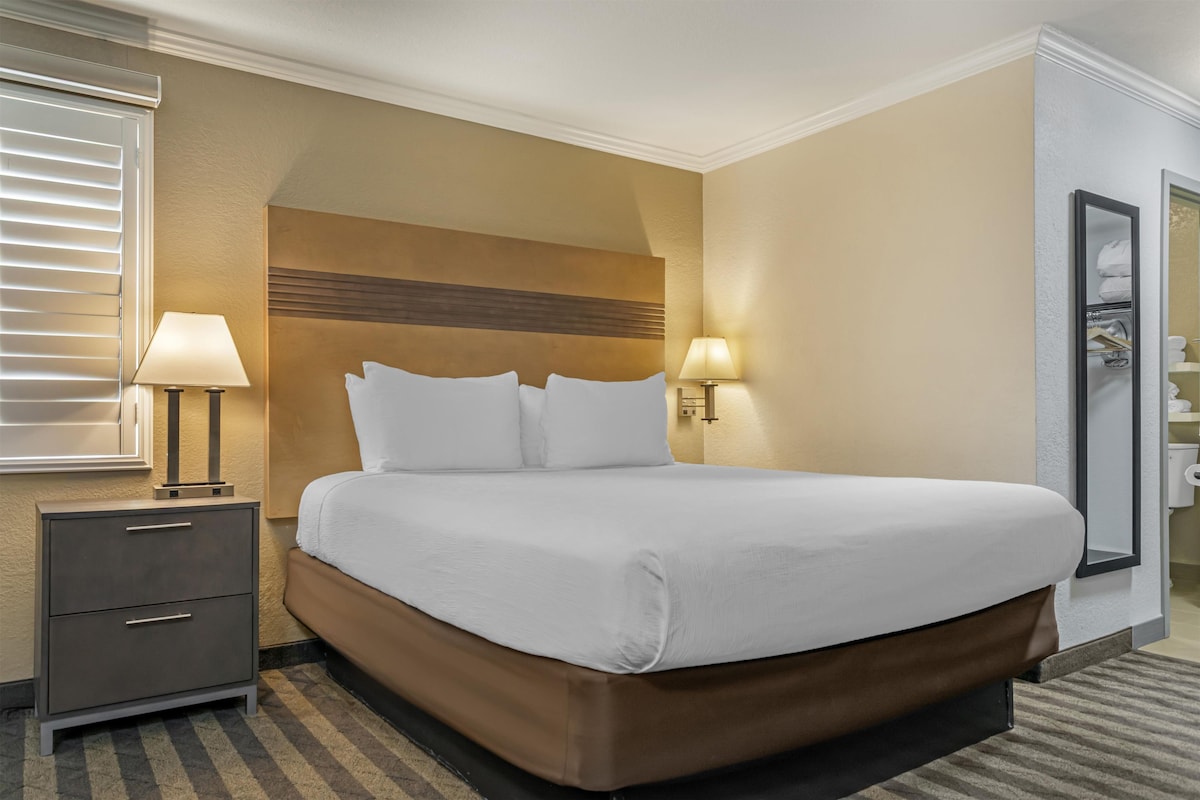 Hotellihuone, jossa on suuri sänky, kaksi yöpöytävalaisinta, yöpöytä laatikoilla ja kylpyhuone, joka näkyy avoimesta ovesta.