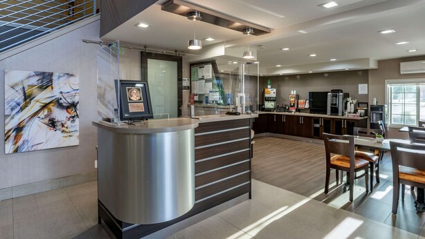Un moderno vestíbulo de hotel con un mostrador de recepción, una escalera, un comedor con sillas de madera y una cocina al fondo.