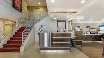 A modern hotel lobby with a staircase, a reception desk, and a kitchenette area.