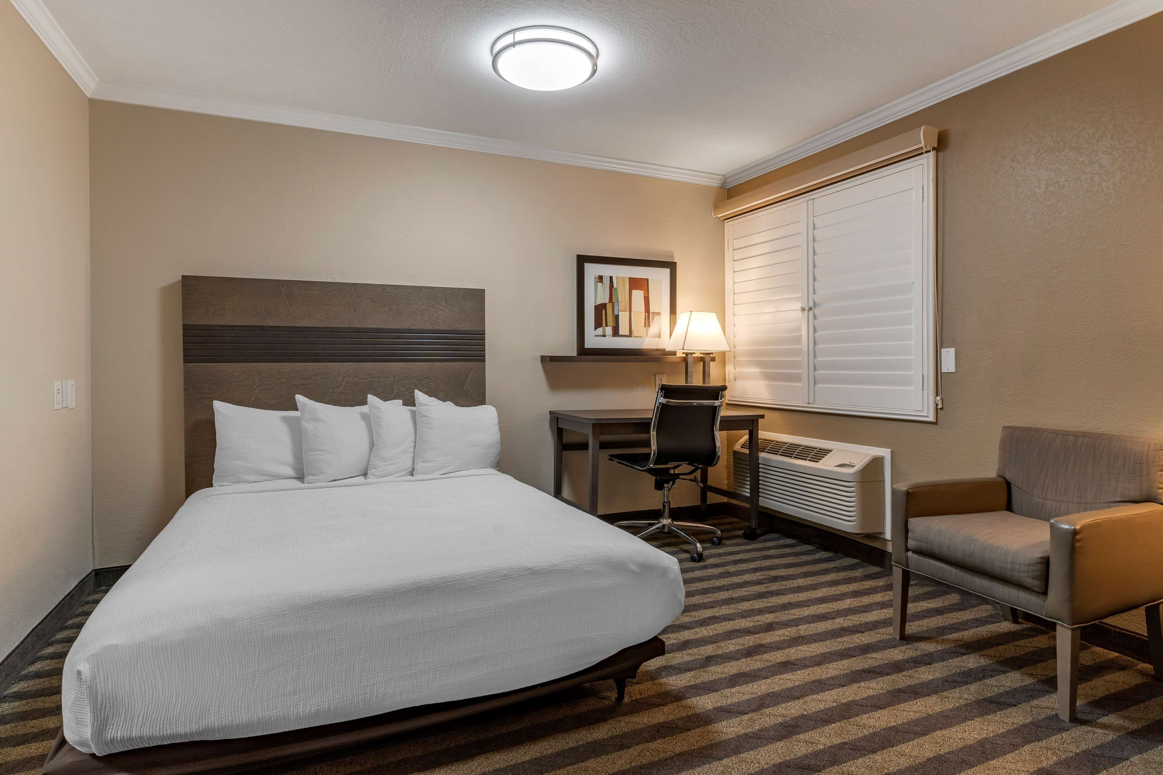 Habitación de hotel con una cama grande, un escritorio con silla, una lámpara y un ventanal con persianas.