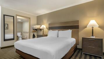 A hotel room with a large bed, a bedside table with a lamp, a mirror, a wardrobe, and a bathroom visible through an open door.