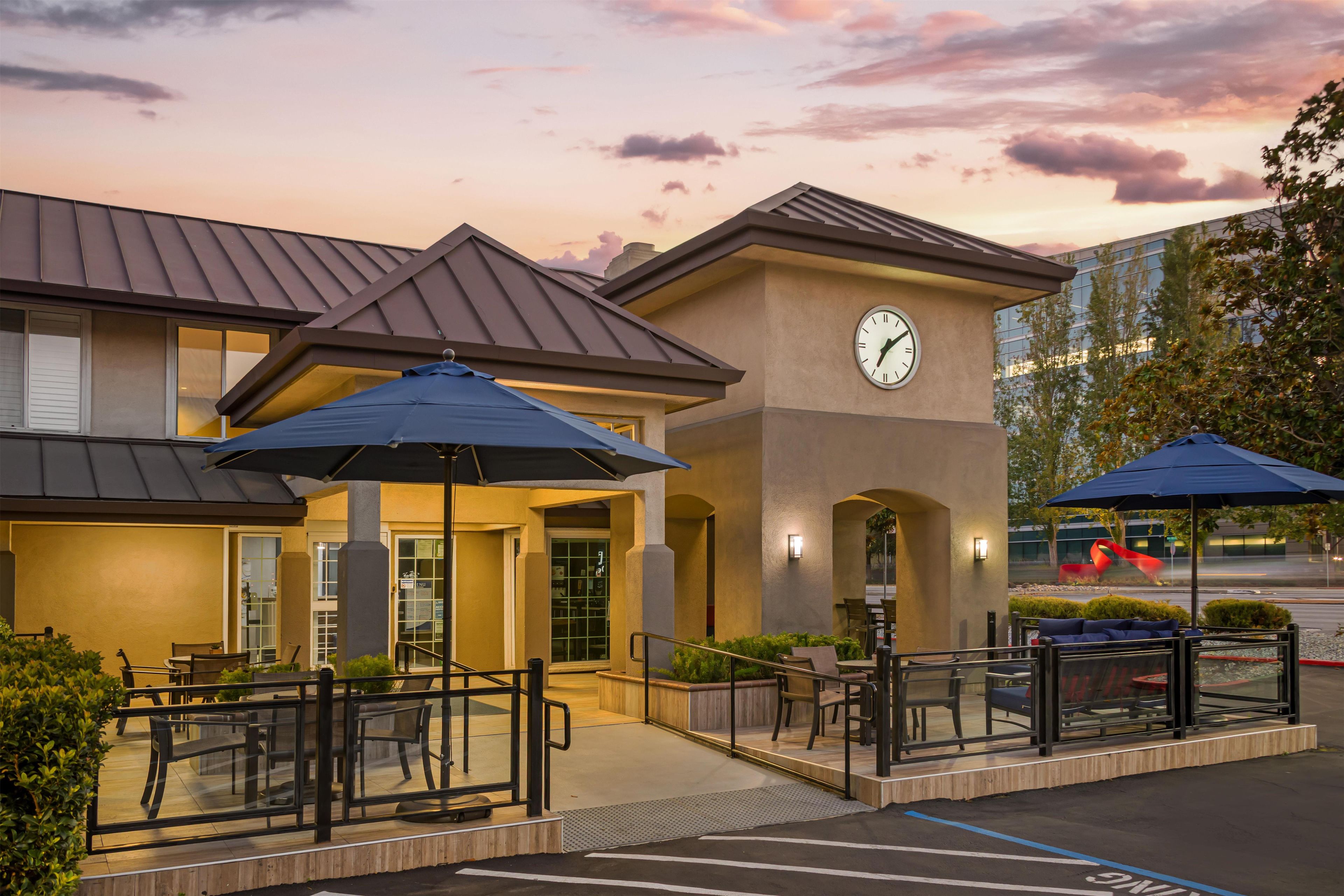 A hotel exterior with outdoor seating, a clock, and a parking lot.
