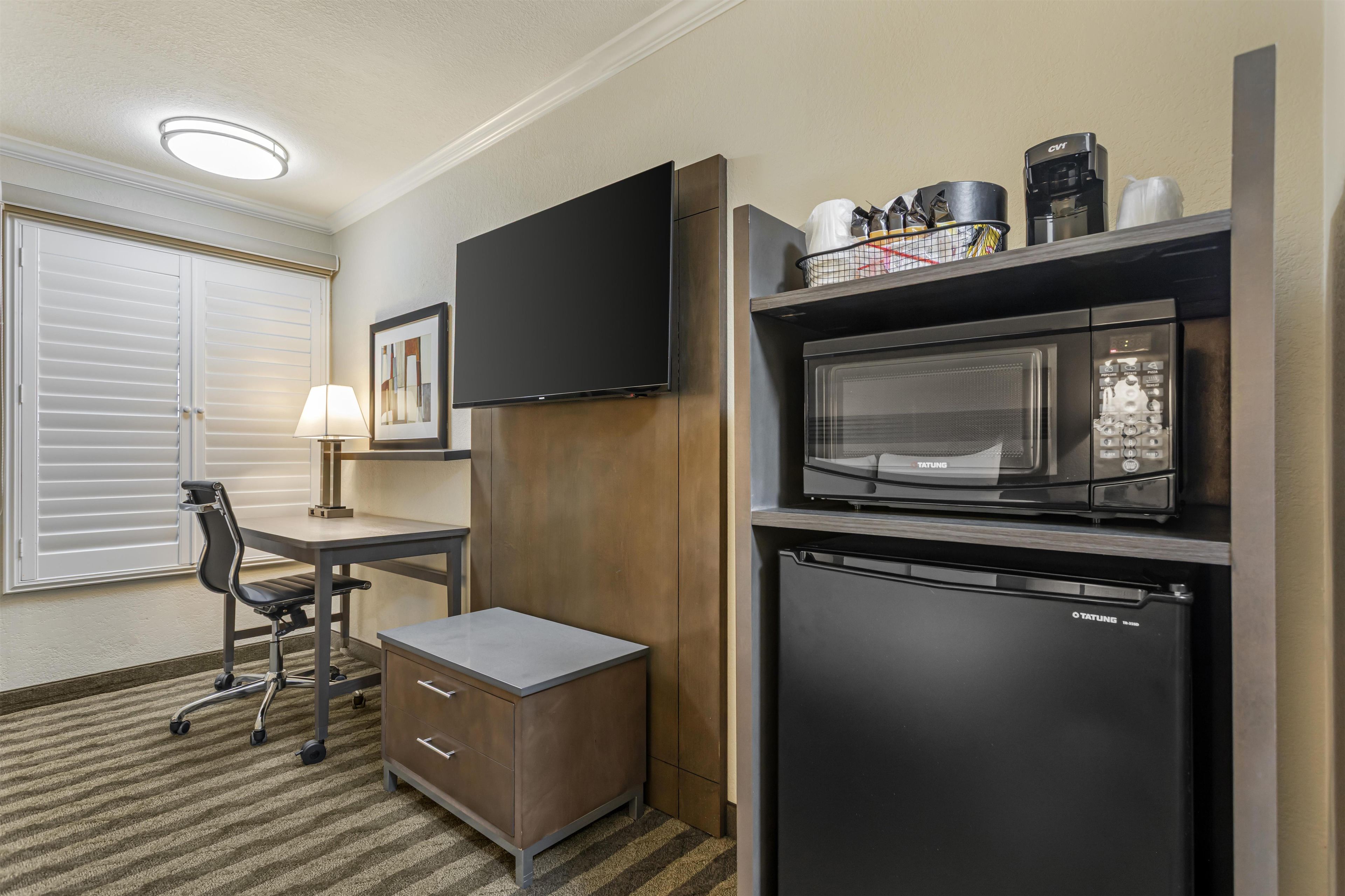 A compact hotel room with a kitchenette, a flat-screen TV mounted on the wall, a small refrigerator, and a desk with a chair.