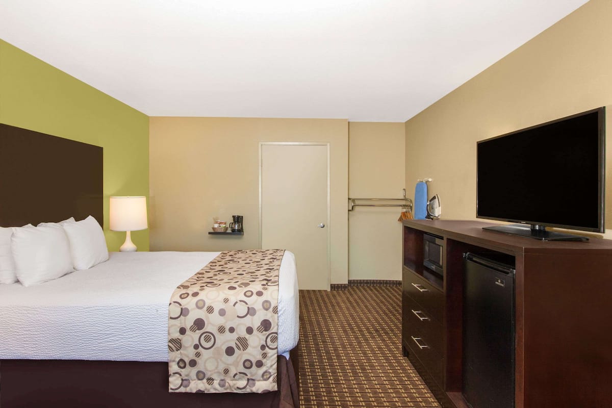 A hotel room with a bed, a television, a small desk with a lamp, and a door.