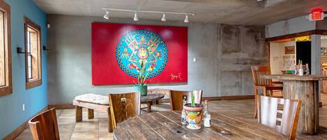 Un espace repas comprenant une table ronde en bois, des chaises, un banc et un grand mur rouge orné d’un motif circulaire coloré.