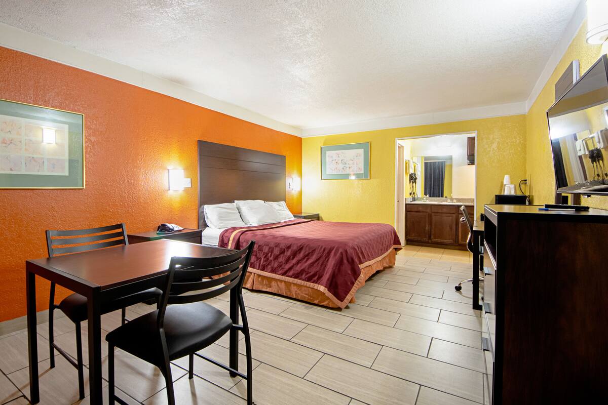 A hotel room with a bed, a desk with chairs, a small kitchenette, and a bathroom visible through an open door.