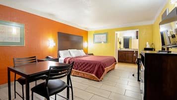 A hotel room with a bed, a desk with chairs, a small kitchenette, and a bathroom visible through an open door.