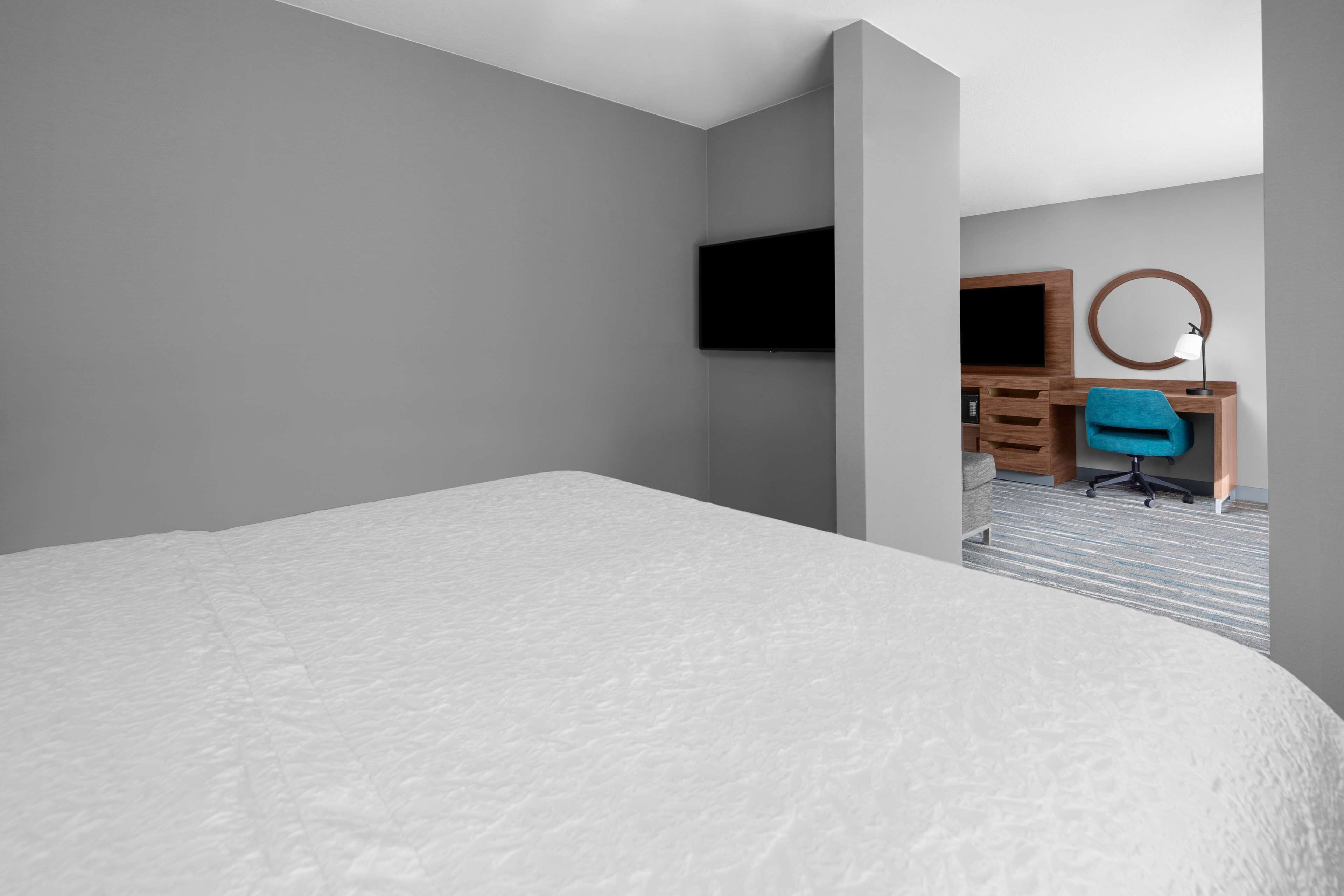 A hotel room with a large bed, a desk with a chair, a TV mounted on the wall, and a round mirror.