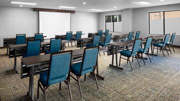 Un salón de conferencias con varias mesas y sillas, una pantalla de proyector y un ventanal grande.