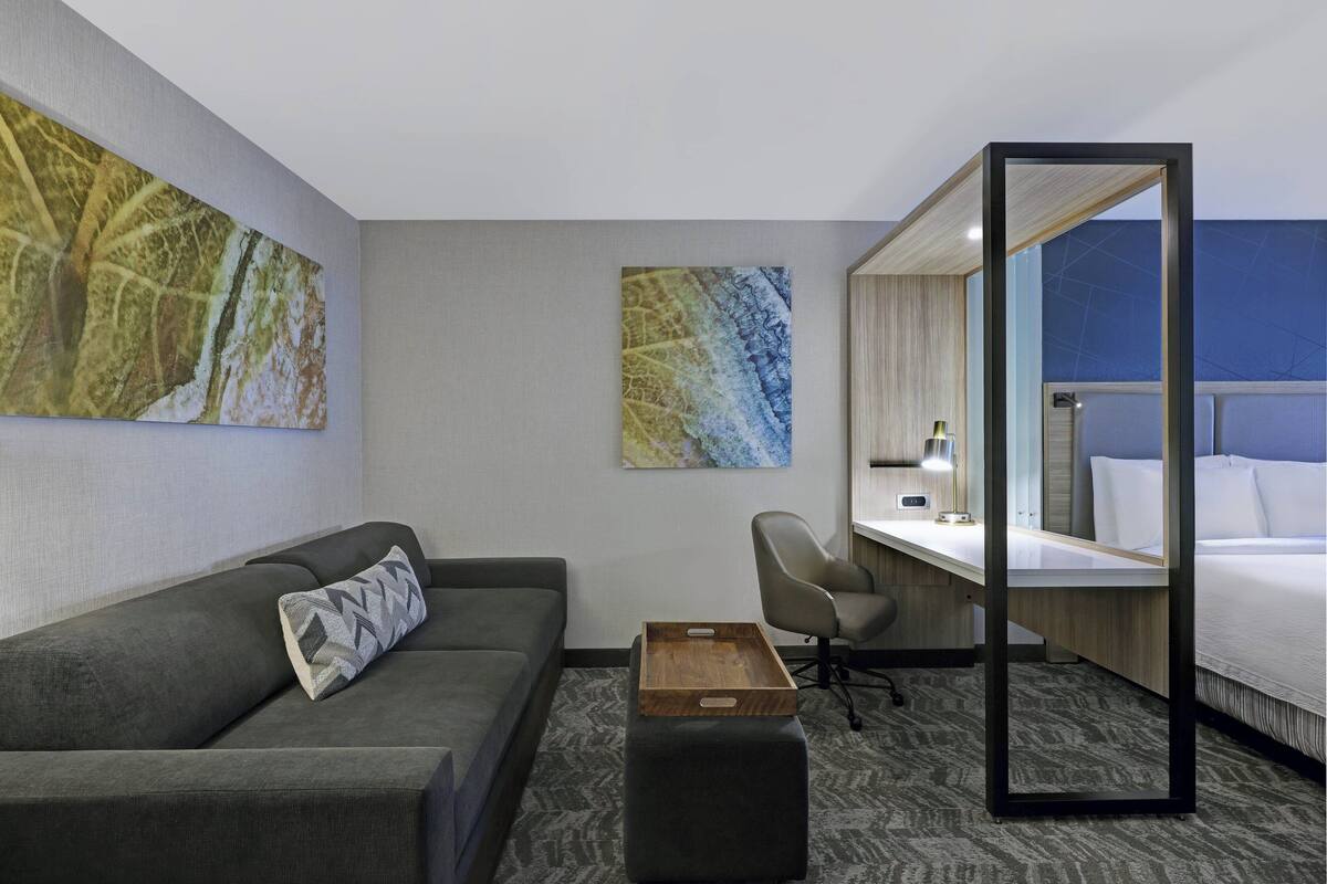 A hotel room with a gray sofa, a wooden side table, a desk with a chair, a bed, and two abstract paintings on the walls.