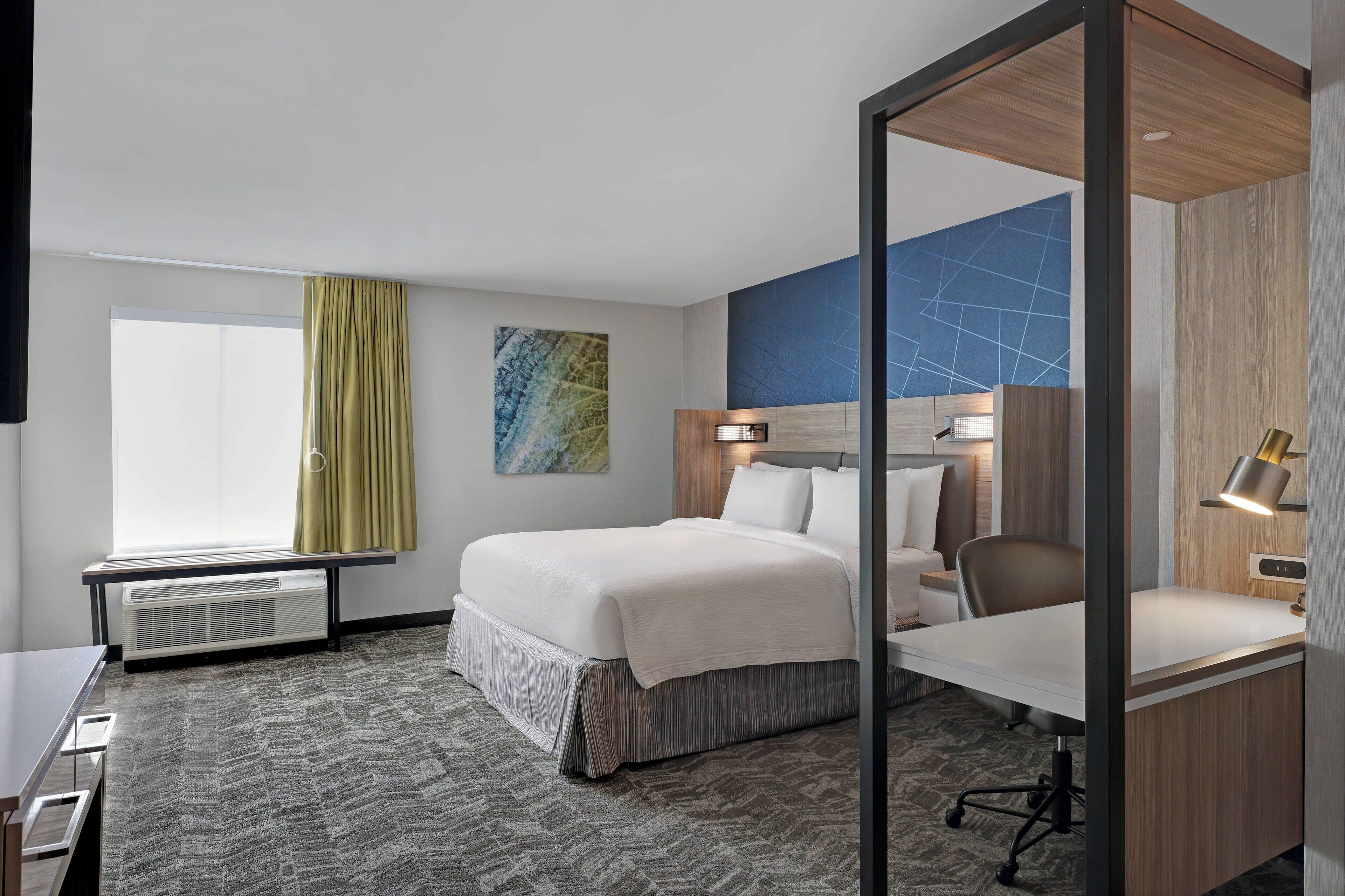 A hotel room with a bed, desk, chair, and a large window with curtains.