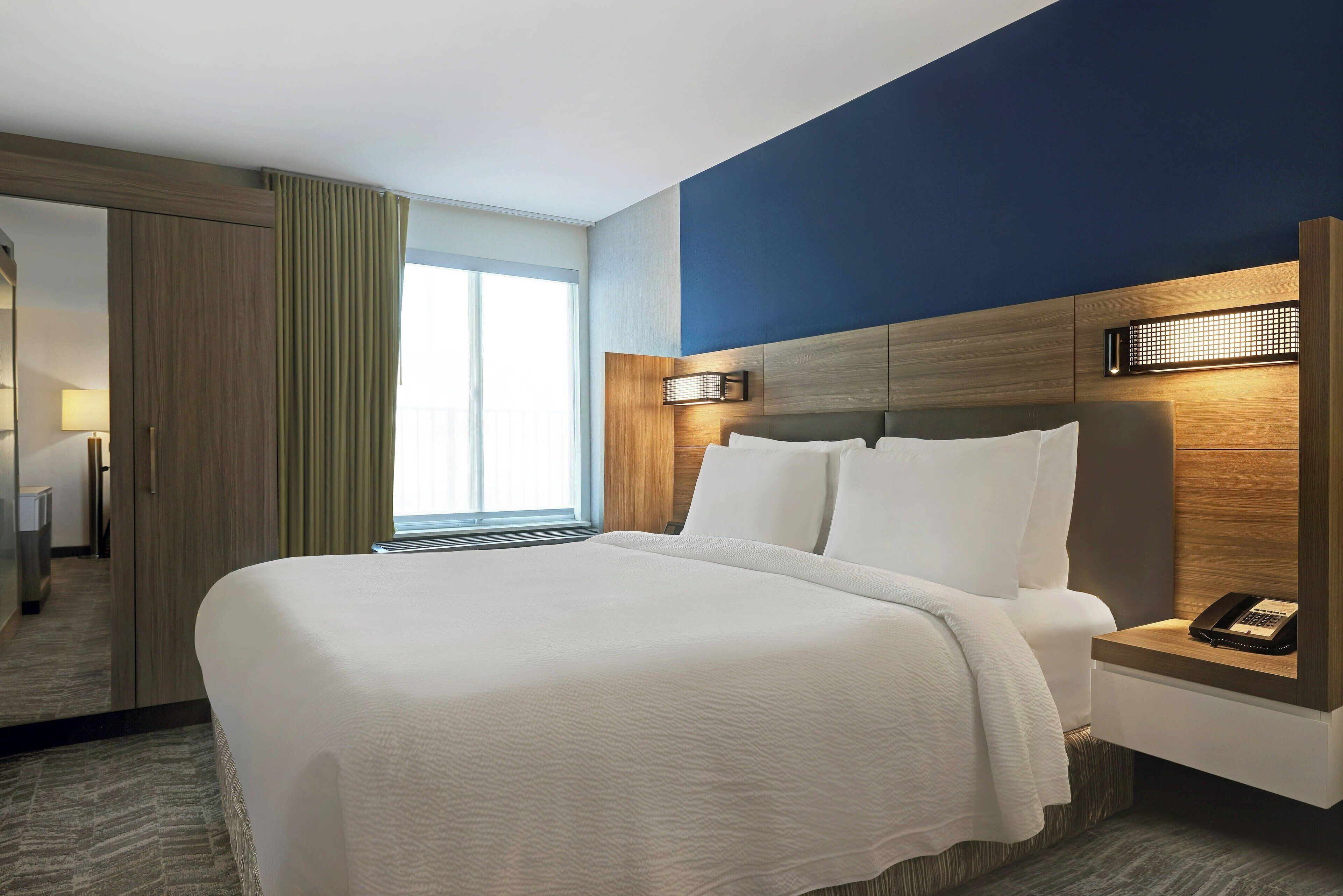 A hotel room with a large bed, a nightstand with a phone, a wall-mounted light, and a view of the hallway.