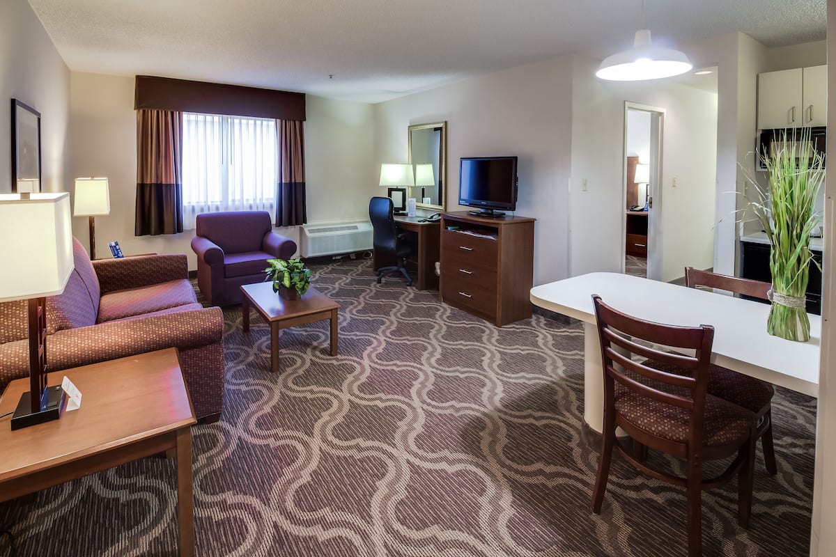 A hotel room with a sofa, armchair, dining table, TV, and a kitchenette area.