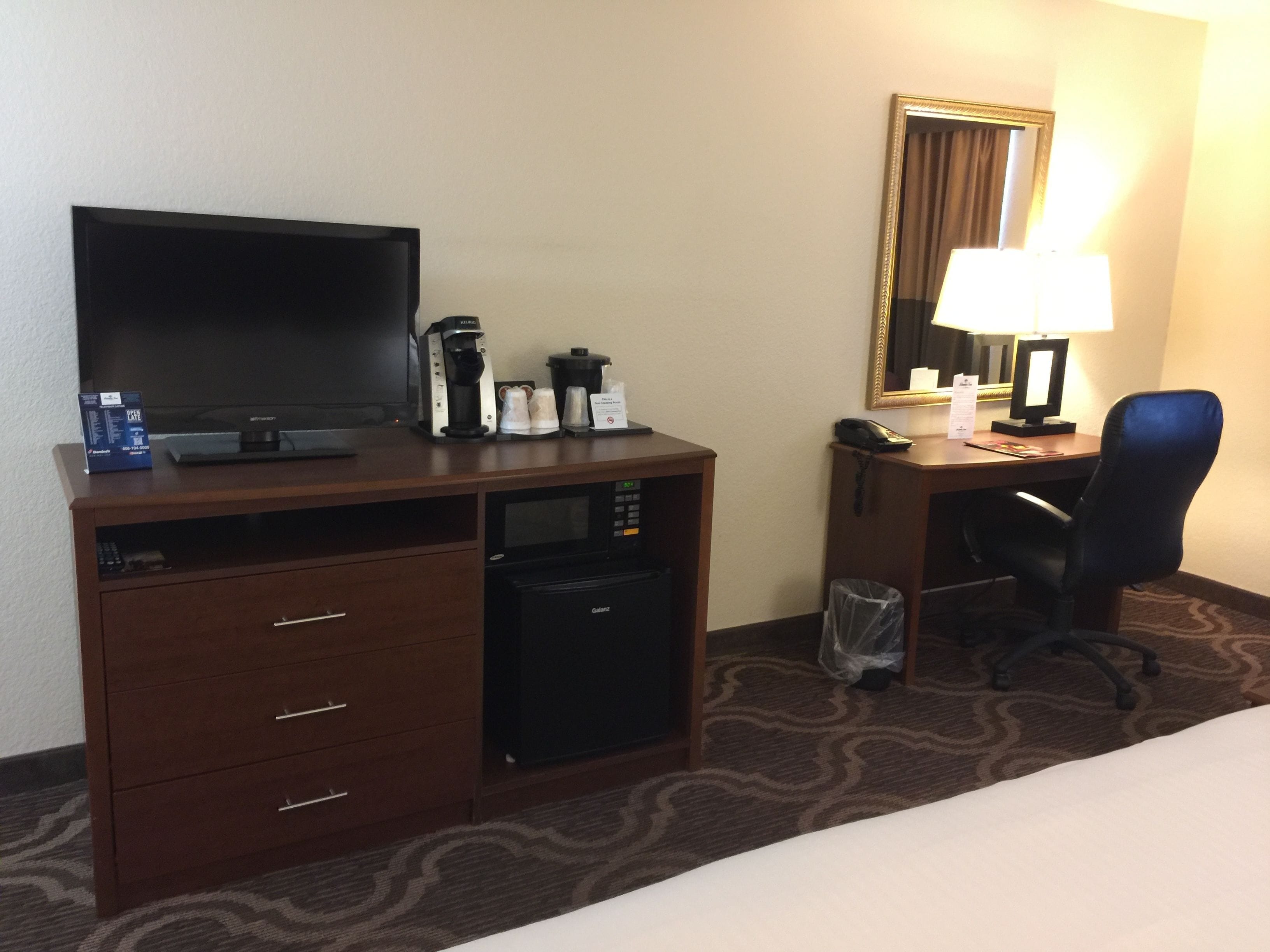 A hotel room with a bed, a desk with a computer, a coffee maker, a TV, a lamp, and a trash can.