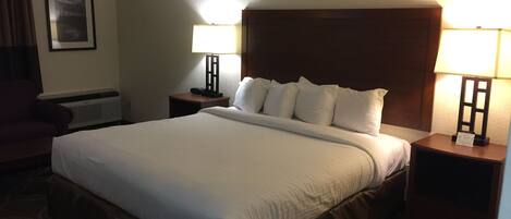 A hotel room with a large bed, two bedside tables with lamps, a chair, a framed picture on the wall, and a wall-mounted heater.