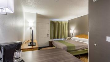 A hotel room with a bed, a desk with a coffee maker, a lamp, and a door.