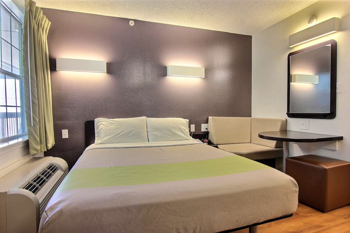 A hotel room with a bed, a sofa, a small table, a mirror, and a window with curtains.