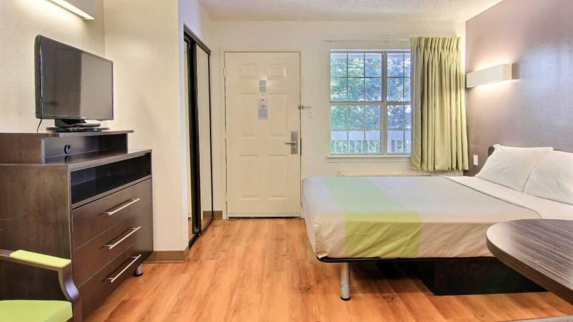 Una habitación de hotel con cama, escritorio con televisor, puerta y ventana con cortinas.