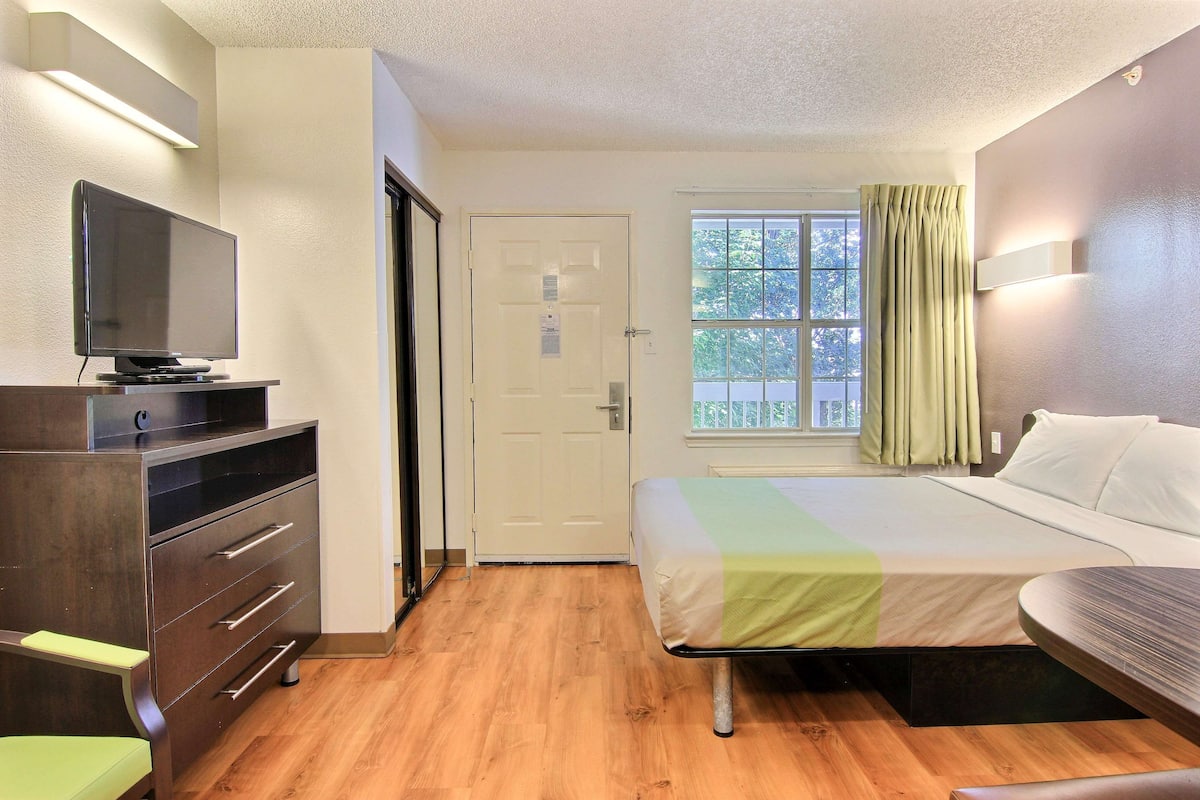 A hotel room with a bed, a TV on a dresser, a window with curtains, and a door.