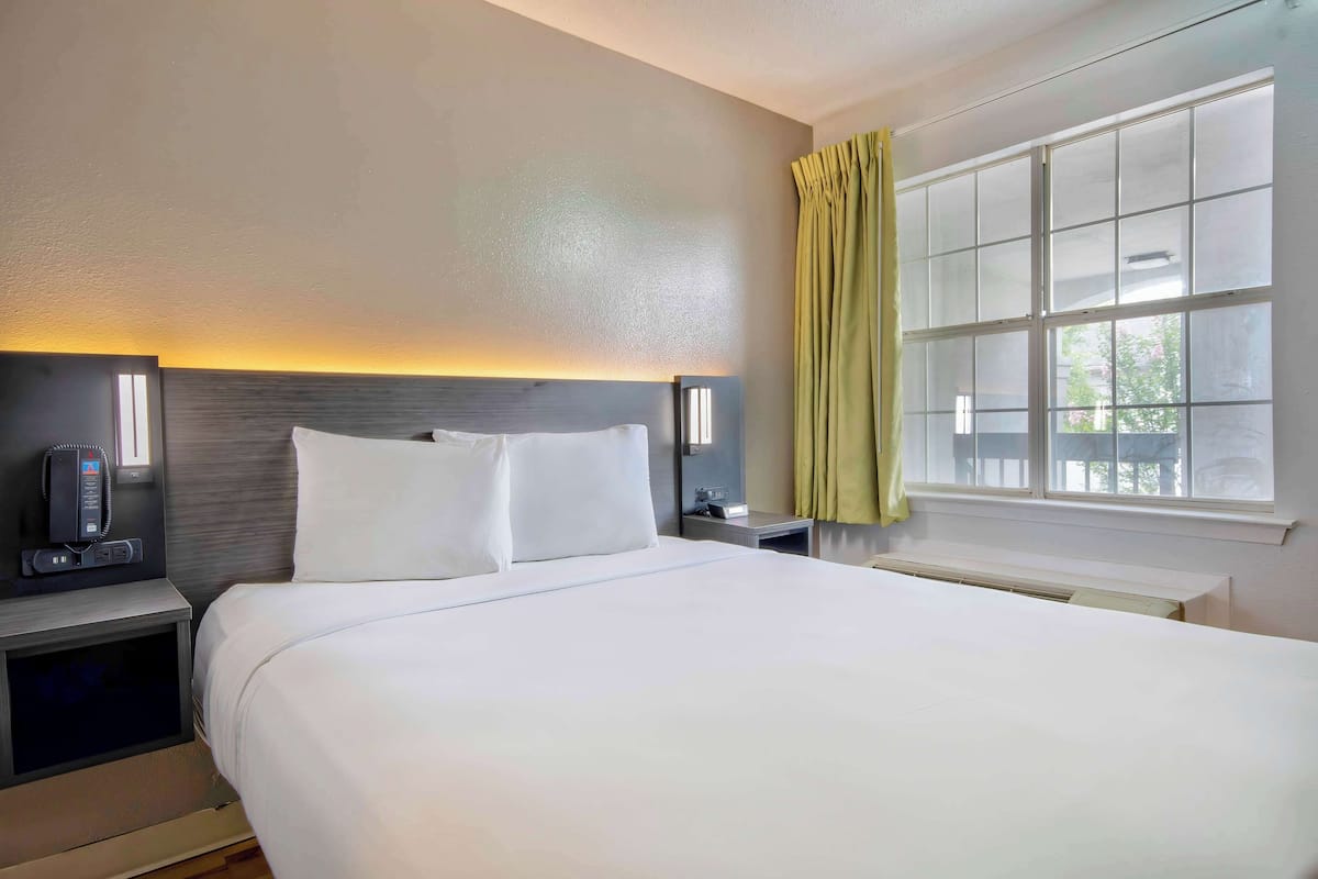 Ein Hotelzimmer mit einem großen Bett, Nachttischen, einem Beistelltisch, einem an der Wand befestigten Telefon und einem Fenster mit Vorhängen.