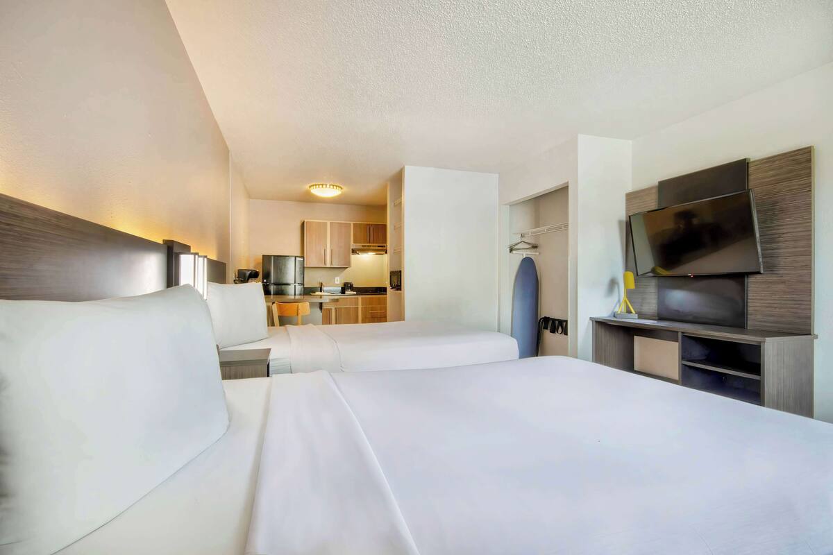 Ein Hotelzimmer mit zwei Betten, einem Fernseher, einem Schreibtisch und einer kleinen Küche im Hintergrund.