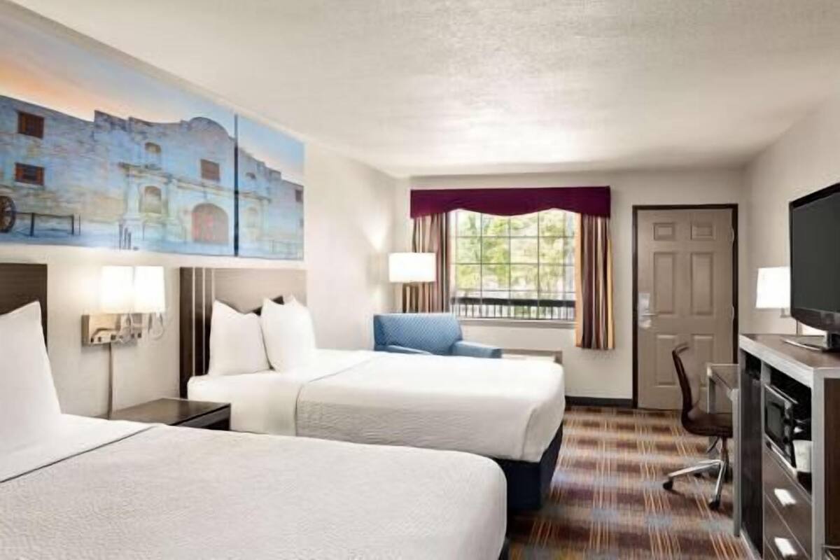 A hotel room with two beds, a desk with a computer, a TV, a window with curtains, and a painting on the wall.