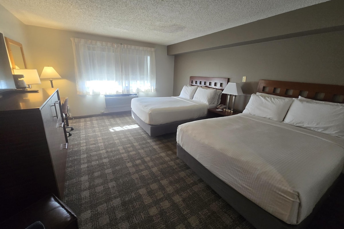 A hotel room with two beds, a desk, a chair, a lamp, and a window with curtains.