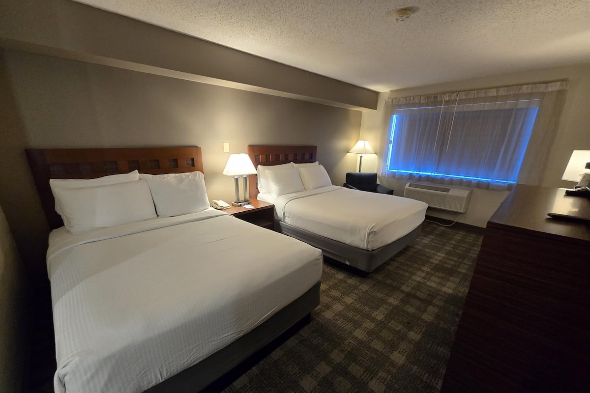 A hotel room with two beds, a desk, a chair, a window with curtains, and a wall-mounted air conditioner.