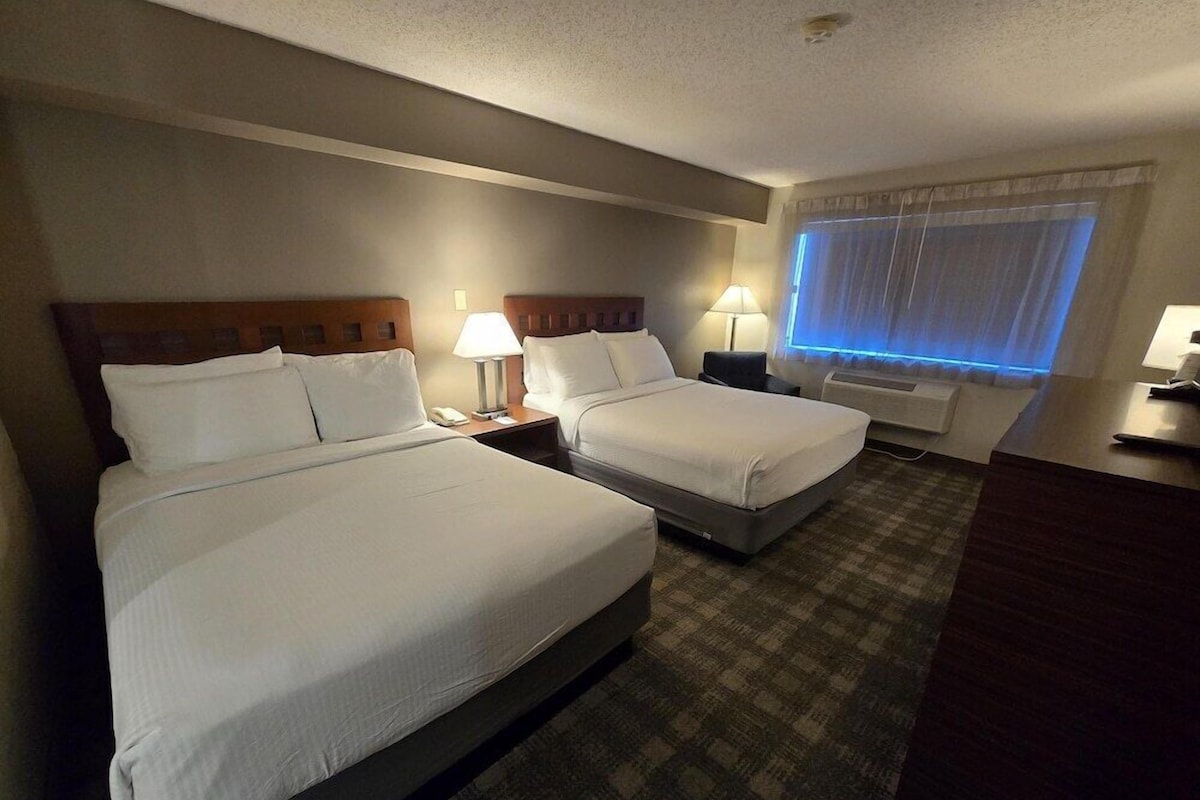 A hotel room with two beds, bedside lamps, a desk, and a window with curtains.