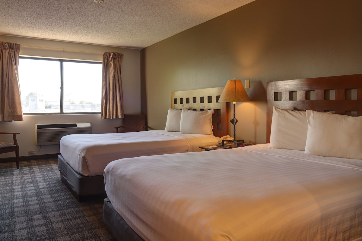 A hotel room with two beds, a desk, a lamp, and a window with curtains.
