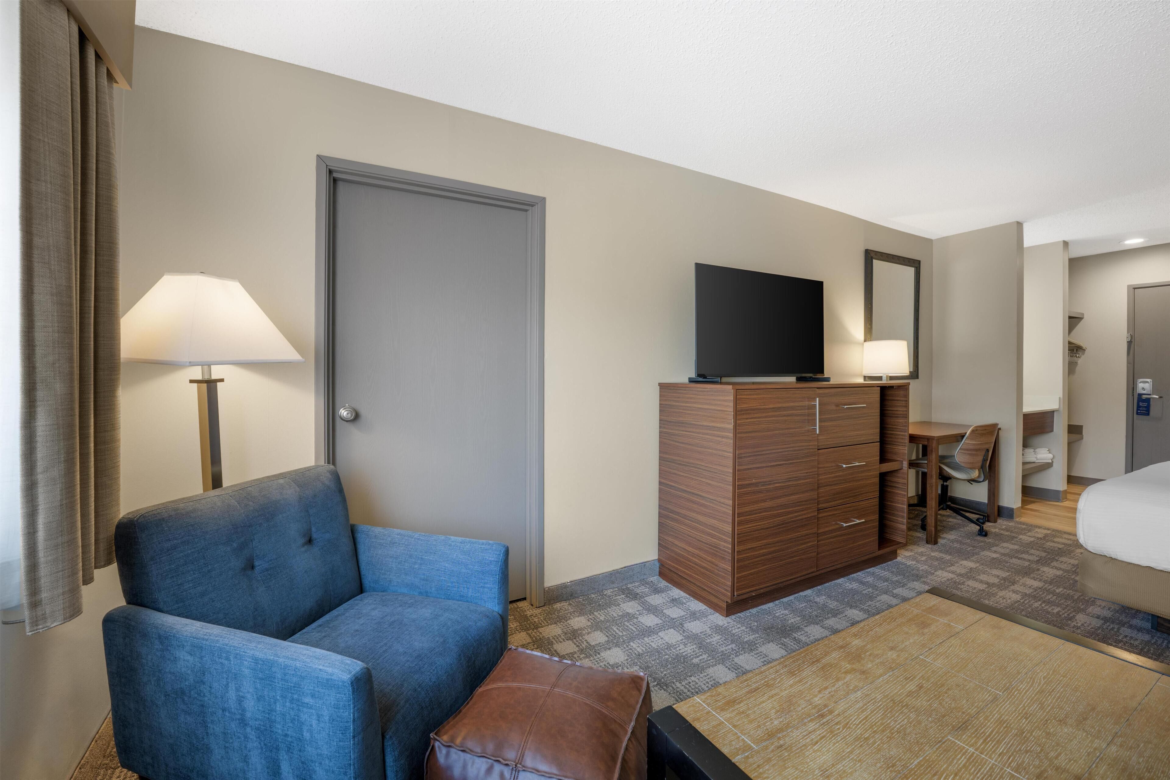 A hotel room with a blue armchair, a wooden dresser, a flat-screen TV, a desk with a chair, and a bed with a white comforter.