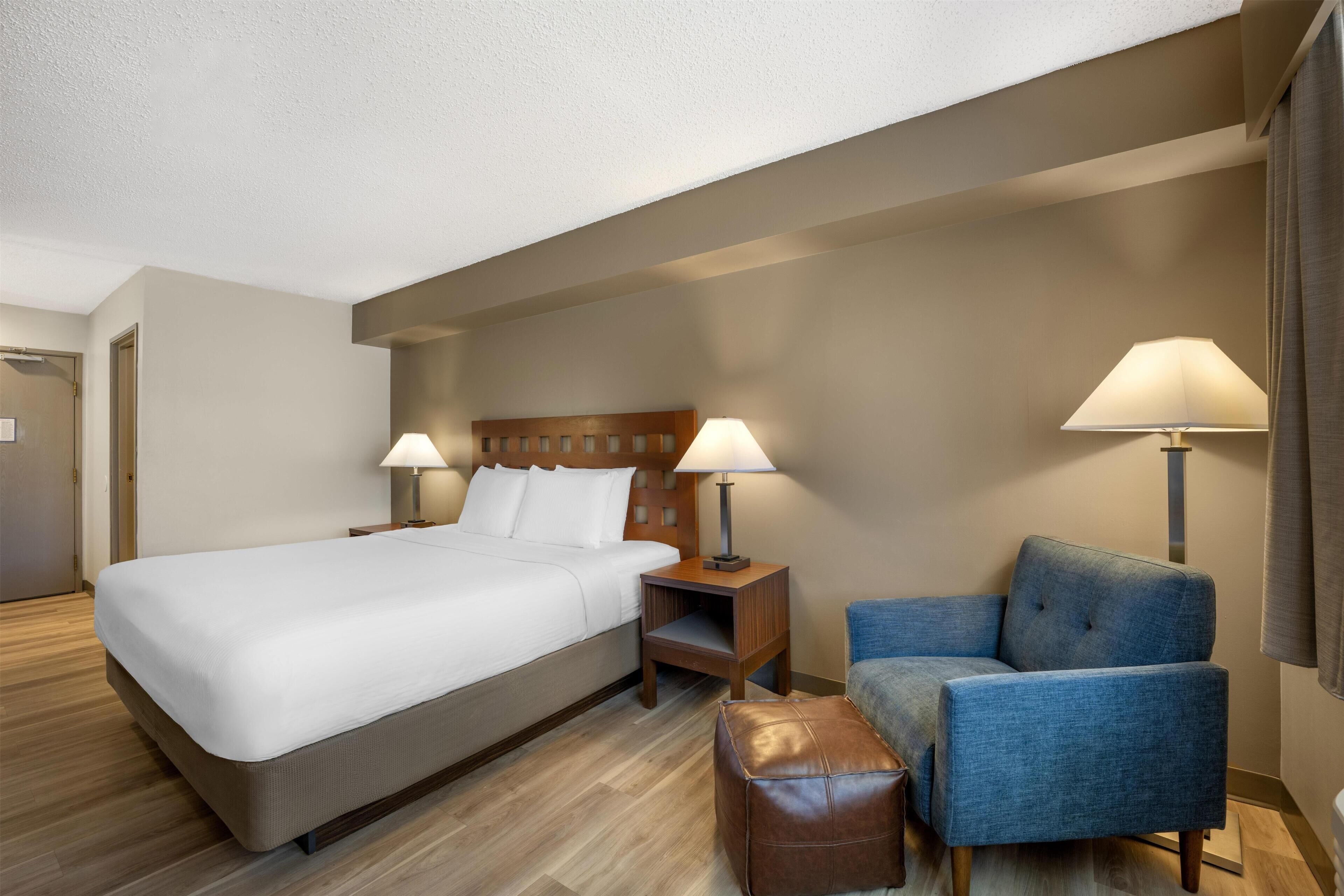 Una habitación de hotel con una cama, dos lámparas de noche, una silla, un pequeño reposapiés y una mesita de noche de madera.