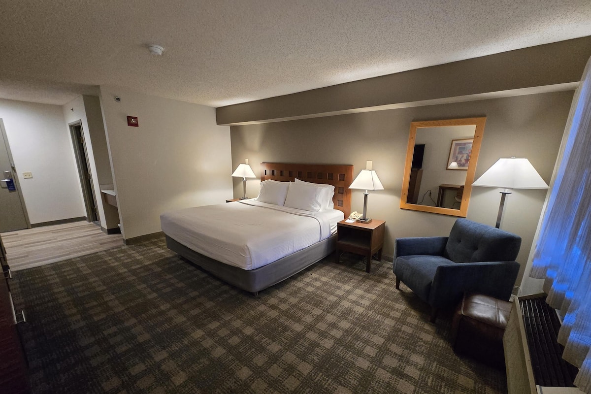 A hotel room with a bed, two bedside tables with lamps, a chair, a mirror, and a carpeted floor.